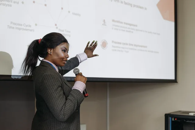 Businesswoman presenting with microphone, discussing improvements with a slide projection in a conference room. Governance für interne Automatisierung