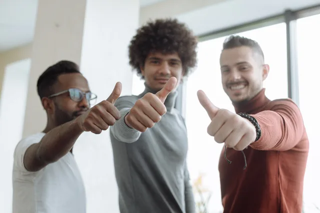 Three diverse men in a team give a thumbs up gesture indoors, symbolizing success and teamwork. Menschliche Freigaben